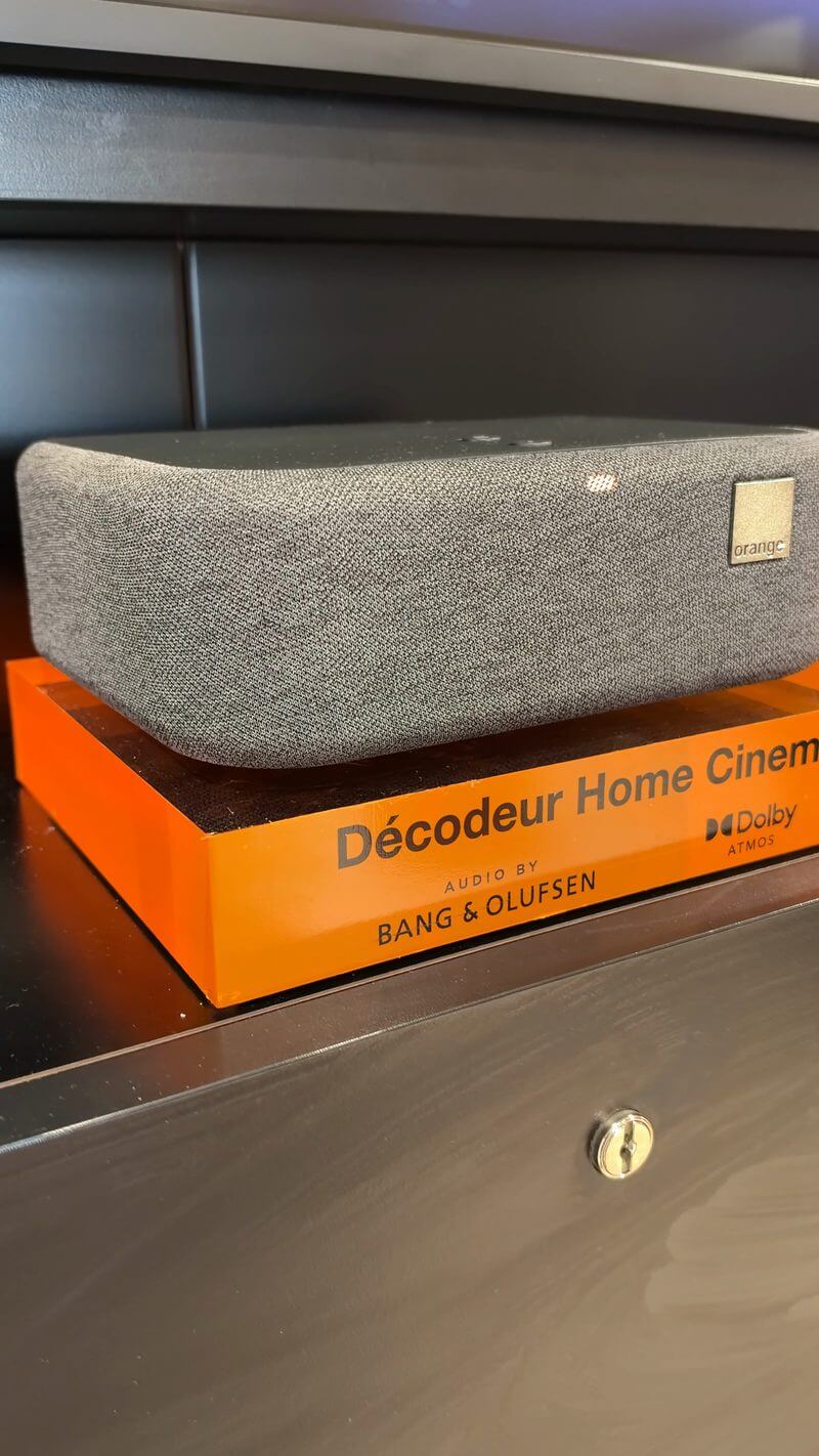 Boîtier gris du Décodeur Home Cinema d'Orange posé sur son socle orange, avec la mention Bang & Olufsen