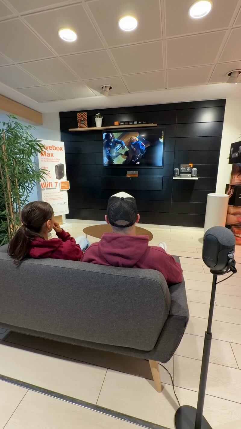 Deux personnes assises sur un canapé face à une télévision pendant la démonstration du Décodeur Home Cinema, une enceinte sur pied à droite