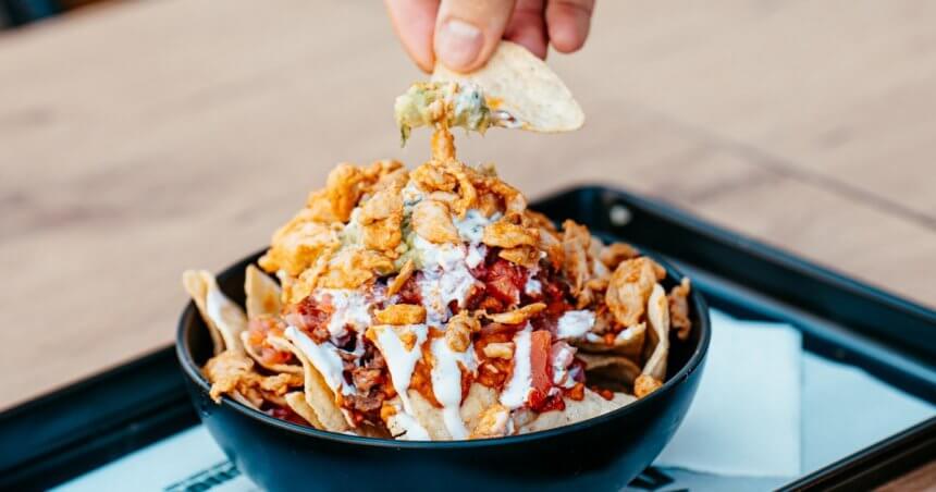 Bol de nachos garnis de viande, tomates, oignons frits et sauce blanche, avec une chips trempée dans le guacamole, chez Nachos à Mérignac